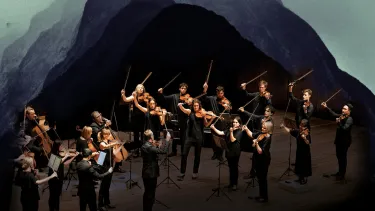 A group of musicians mid-performance on a stage collaged into the side of a mountain with dark mist and clouds in the background.