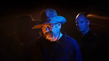 Mark Atkins is in a cowboy hat is lit with dramatic blue and orange lighting against a dark background. Erkki Veltheim stands blurred behind him, creating a mysterious tone.