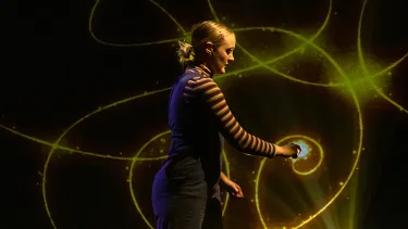 A person in a striped shirt and overalls draws a gold sparkling line on a black background.