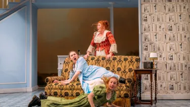 Three cast members position themselves on three levels around a yellow couch.