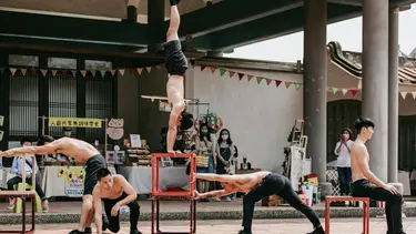 A man does a handstand on top of two scaffolding like cubes. On either side various other performers are doing stretches or poses.