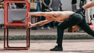 A performer is leant over and outstretched grabbing on to two cubes. A foot of another performer is visible on his back.