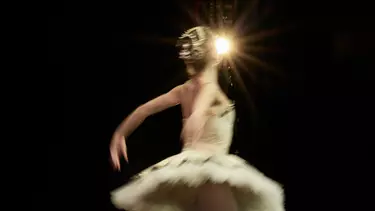 A ballerina wears a gold tutu and hairpiece. Her back is to the camera and her arms are stretched behind her, mid-dance move. A spotlight is seen behind her.