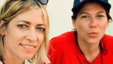 A blonde woman with glasses on her head sits next to another women wearing a hat and a red wutang shirt. They have festival lanyards on.