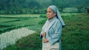 A Nun wearing a light blue habit overlooks a green field while clutching a white bag.