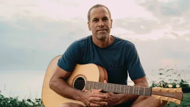 A man sits cross legged on grass. He is holding a guitar and wearing a blue t-shirt.
