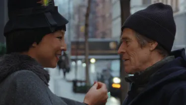 A woman wearing a coat and a hat with a feather in it stands on an urban street facing a man who is wearing a beanie and coat.