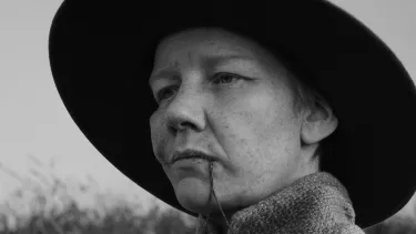 A person is looking off into the distance in a black and white image. They are wearing a wide brimmed hat and have a piece of straw in their mouth.