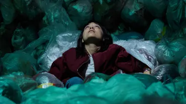 A person in a grey shirt and red jacket is looking upward while seated on and surrounded by full garbage bags.