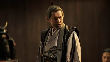 A man in a traditional Japanese garment is standing while other men dressed similarly are  crouched and surrounding him. A helmet and armour is visible in the background.