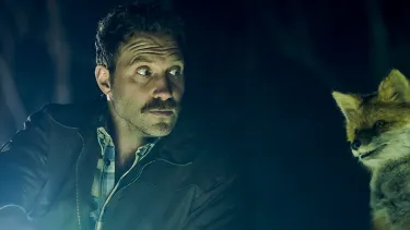 A moustached man is crouched next to an animatronic fox. He is holding a flash light as it is night time in the woods.