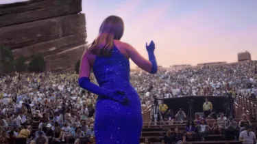 A woman in a blue dress and gloves waves to a crowd from on top of a stage. In the background is a large rock or cliff.