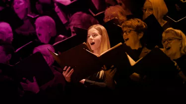 A blonde girl with an open song book sings amongst a choir