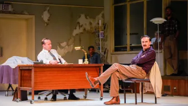 Man sits down with arms folded across a man sitting at a desk.