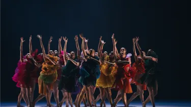 A large group of dancers dressed in a range of pinks and orange hold one arm aloft.