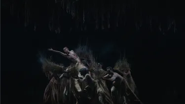A lone dancer kneels on a group of dancers surrounding him in low light.