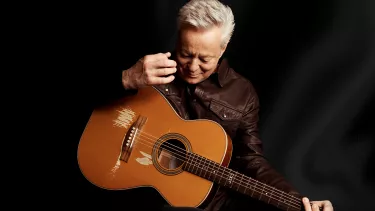 A man with grey hairs wearing a black leather jacket playing a guitar.