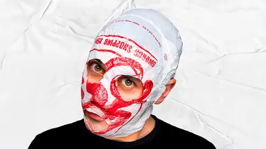 A man in front of a white textured background is wearing a black shirt and a plastic bag over his head. The white bag with red design has eye holes and a mouth hole cut out.