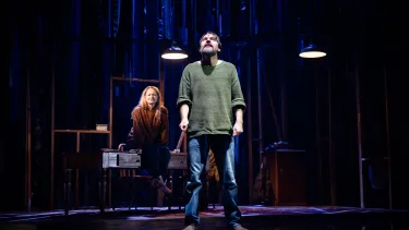 In the foreground a man in a green sweater looks upwards into a light. In the background a red haired woman sits on top of a desk.