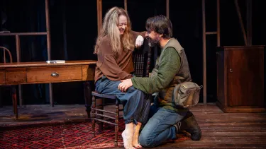 A woman sits on a chair while a man kneels looking up at her with his hand on her knee.
