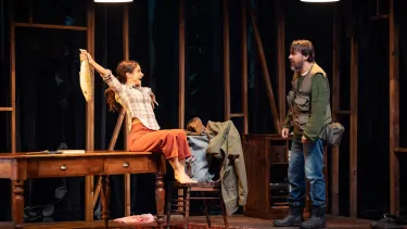 A woman in a plaid shirt and orange pants sits on top of a table with her arms out stretched. She is holding a fish in one hand. A man looks on in shock.