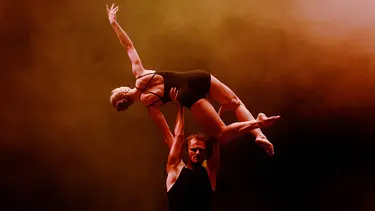 A male dancer lifts a female dancer in a black leotard gracefully overhead against a dramatic orange-lit background.