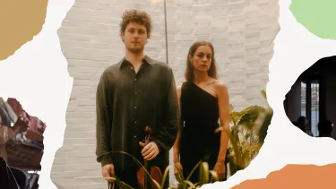 A man and woman stand side by side in a bright room, surrounded by plants. The man holds a violin.