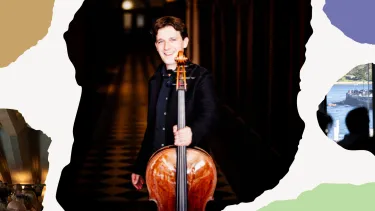 A white man smiles while holding a cello in a dimly lit hallway.