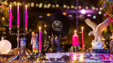 A vibrant table with flowers, a vinyl record, pink candles, and decorative items. The Sydney Harbour Bridge can be seen in the distance at night.