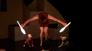 A man is leaning over holding two glowing juggling sticks. Two young children watch on from either side.