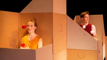 A man and a woman stand inside a box structure both juggling balls.