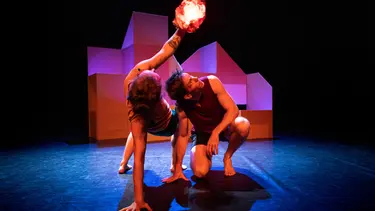 Two performers are crouched to the ground. One is leaning back holding up something that resembles a glowing fire.