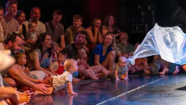 The front row of a crowd is visible. Parents and young children look towards a stage where a floating white fabric is visible.