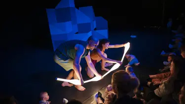 Two performers hold up four glowing sticks on a dimly lit stage.