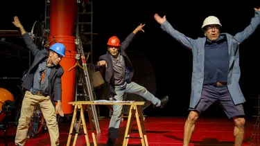 Three men in different coloured hardhats dance on a set resembling a construction site.