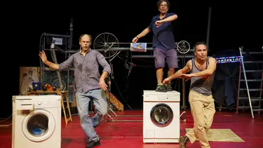 Three men dance while fixing washing machines. Two are by their side while the third is on top of a washing machine.