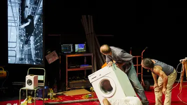 Three men are looking at a screen while attempting to repair a washing machine. One man is on the ground looking underneath it.