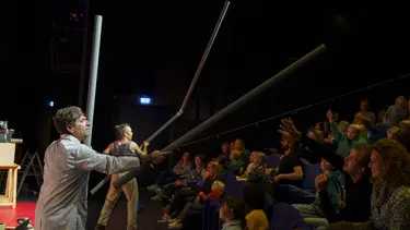 Two performers hold out pipes towards a crowd of people in tiered seating.