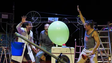 Two men are theatrically holding their hands above a red button and large green balloon. Another looks on with a shocked look.