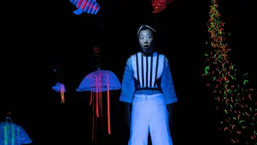 A performer wearing fluorescent white clothing against a black background is surrounded by glow in the dark sea creatures made out of various materials.