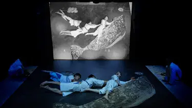 Three performers lay on the floor holding on to each others backs. They are holding onto the shape of a whale. Their images are projected from above onto a screen.