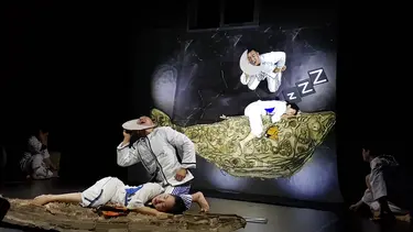 A performer lays asleep on the cutout of a whale. Another performer holds a cardboard cutout of the crescent moon to his face.