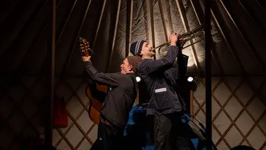 Two men stand back to back inside of a tent. They are wearing jackets and beanies, one is playing a trumpet and the other a guitar.