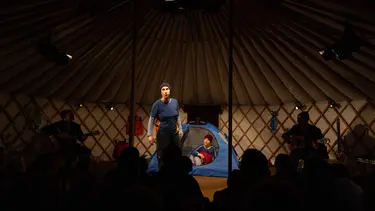 Three performers are on stage in front of a crowd. One sits in a tent, one is in the foreground and the others are off to the side playing guitars.