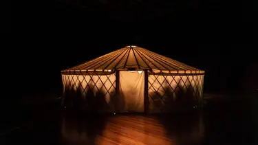 A large canvas tent sits on a stage. It is lit up on the inside making the silhouettes of a group of people visible.