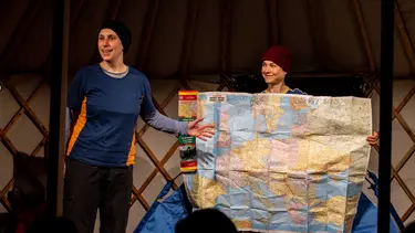 Two performers stand onstage in beanies and warm tops. One is holding up a large map while the other gestures toward it.