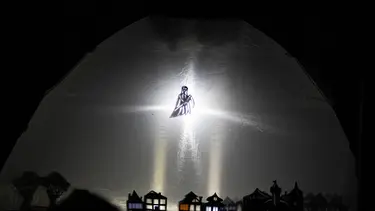 The inside of a tent is lit making shaddow puppetry of a town and grim reaper visible.