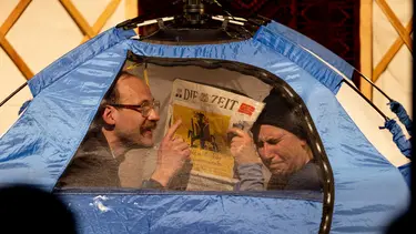 Two performers sit inside a tent. One is holding a newspaper crying while the other is wearing glasses and a moustache and is pointing.