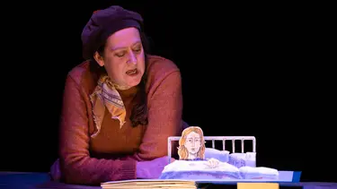 A woman in a beret and orange sweater leans over a table which contains a fold-out picture book.