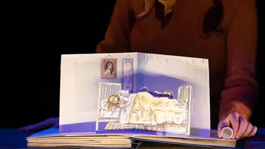 A fold up picture book is on a table. It displays and image of a man and woman sleeping top to tail in a bed.